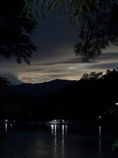 The image depicts a nighttime scene at the Mekong River in Choumkhong, Laos. Silhouetted against a dimly lit sky with striated clouds are dark, undulating mountains. In the foreground, the calm river surface reflects the lights of a floating structure with multiple illuminated sections, possibly a restaurant or resort. The water also shows reflections of distant lights and the faint shimmer of the moon. Trees and foliage frame the top and sides of the image, adding to the sense of depth and seclusion. The overall atmosphere is serene and peaceful, with the natural landscape dominated by darkness and illuminated by man-made lights.