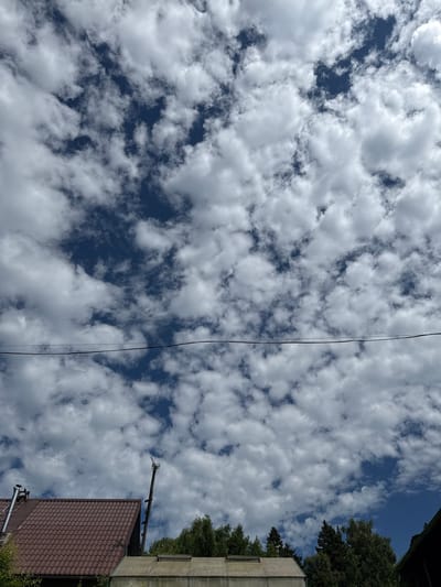The image captures a bright, partly cloudy sky over a rural or suburban area. The sky is filled with scattered white cumulus clouds against a deep blue background, suggesting fair weather. A dark, thin wire stretches horizontally across the mid-section of the frame, likely a power line or telephone wire.
In the lower portion of the image, the rooftops of several structures are visible. On the left, a brown, tiled roof of a house or building dominates the foreground, with a metal chimney pipe extending from its peak. Adjacent to it, to the right, is a lighter colored, flat-roofed structure, possibly a greenhouse or shed, with translucent panels. A tall, dark pole with what appears to be an antenna or communication device is erected behind the brown roof, reaching into the sky.
A dense line of green trees forms a backdrop behind the structures, with various species of trees visible. The overall environment appears to be a residential area or a garden community, consistent with a location like Дзержинец-83 Снт, Russia. No people are present in the frame. The lighting suggests it is daytime, likely late morning or early afternoon, given the brightness of the clouds and the clear sky.