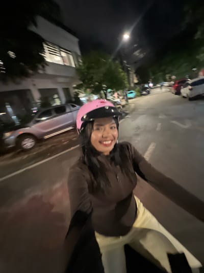 This is a first-person perspective, night time, selfie-style photo taken from a moving vehicle. A person wearing a brown zip-up jacket and light-colored pants is visible. They are wearing a pink helmet with spikes on top and have dark hair. They are smiling directly at the camera. In the background, a street is visible with parked cars, trees, and buildings. The scene appears to be in a residential or commercial area with streetlights and the glow from building windows. The location is Minh Xuân Ward, Vietnam.
