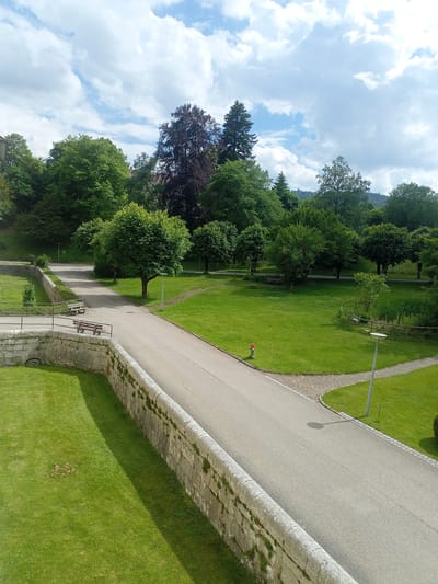 This media file captures an outdoor scene on a partly cloudy day. The primary subjects are the lush green landscape, a paved pathway, and a stone wall. The pathway curves through a well-maintained park with green grass and scattered trees. On the pathway, there are two wooden benches visible, suggesting a place for rest and contemplation. A fire hydrant is also present near the edge of the path. The stone wall, constructed with large, rough-hewn blocks, runs along the foreground and appears to be an old fortification or retaining wall. Beyond the immediate park area, a dense forest of trees can be seen, with some taller, darker trees standing out. The sky is a mix of blue sky and fluffy white clouds, indicating fair weather. The lighting suggests it is daytime, likely late morning or early afternoon, given the shadows cast by the trees and the light intensity. There is no text visible in the image. The overall impression is one of a serene and picturesque park setting, possibly in a historical or natural area.