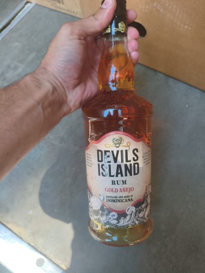 A person's hand holds a bottle of "Devil's Island Rum Gold Añejo" up to the camera. The rum is amber in color, and the bottle has a dark cap and a label featuring a skull and crossbones motif, as well as ornate scrollwork. The label also indicates that the rum is "Distilled and aged in Dominicana." In the background, a portion of a cardboard box is visible, along with a textured surface that appears to be outdoors. The lighting suggests it is daytime, and there is a shadow cast by the bottle and hand.