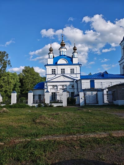 The image depicts a Russian Orthodox church with white walls and blue roofs and domes. The church has several golden spires. A black wrought-iron fence surrounds the church property, with grass in the foreground. Trees are visible behind the church, and a blue sky with fluffy white clouds provides the backdrop. The image was taken in Мамонтово, Russia.
