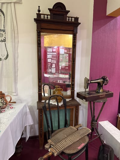 The image depicts an interior scene, likely a museum or a historical exhibit in Fort-Shevchenko, Kazakhstan. In the center of the frame, a dark wooden hall tree with a mirror stands against a pale wall. The mirror reflects a display behind it, featuring framed photographs and objects. To the left of the hall tree, a table is covered with a white tablecloth adorned with lace and embroidered flowers. On the table, a decorative ceramic teapot with floral motifs is visible.

To the right of the hall tree, a vintage black sewing machine with gold embellishments rests on a wooden base, which is mounted on a rustic, tripod-like stand. This stand is placed against a vibrant magenta wall. In the foreground, a wooden chair with a woven seat holds two wooden washboards. The floor is covered with a dark red carpet. The overall impression is one of an organized display of historical artifacts, likely related to domestic life and crafts.