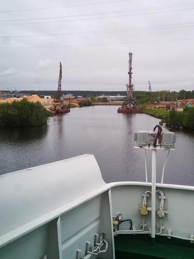 The image depicts a view from a ship sailing on a river towards what appears to be a lumber mill or storage facility in Depo, Russia. The sky is overcast, creating a muted light. Two large cranes are visible on either side of the riverbank, with piles of lumber and other industrial materials nearby. The river's water is dark, reflecting the sky. The ship's white exterior is prominently featured in the foreground, including some of its mechanical and electrical components.