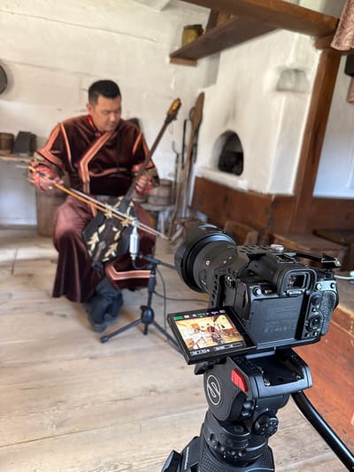 In a rustic room with whitewashed walls and wooden floorboards, a man dressed in traditional clothing, including a brown embroidered robe, is seated and playing a stringed instrument resembling a fiddle. He is focused on his playing, holding a bow in his right hand and the instrument in his left. In the foreground, a professional camera on a tripod is positioned to film the musician. The camera's screen shows a live feed of the performance, indicating a recording is in progress. The background reveals a traditional Russian stove and other rustic elements, suggesting a historical or cultural setting, possibly in the city of Shushenskoye, Russia, known for its historical significance and ethnographic museums.
