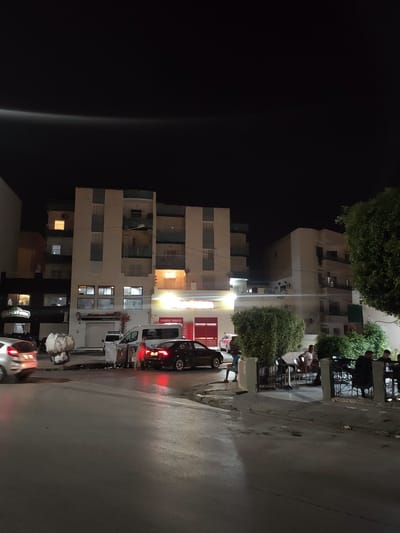 This is a nighttime exterior photograph of a street scene in an urban environment. The scene is dominated by the facade of a multi-story apartment building with balconies and lit windows. In front of the building, there is a street with several vehicles, including a silver hatchback in motion on the left, a white van, and a black sedan. Adjacent to the vehicles are trash receptacles and a figure walking with a cart. To the right of the apartment building, there is a sidewalk area with several people seated at tables outside a establishment, possibly a cafe or restaurant, bordered by a metal fence and lush green bushes. The sky above is dark. The overall impression is a typical street view at night in a residential and commercial area.