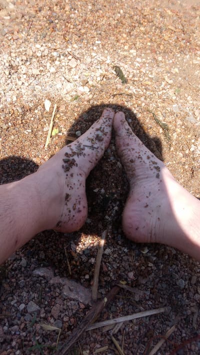 A close-up overhead shot shows two bare feet positioned with the toes touching and the soles facing upwards, partially submerged in coarse, damp, brown gravel. Small pieces of debris, including twigs and plant matter, are adhering to the skin of the feet. The gravel is a mix of small, rounded stones of varying shades of brown and grey. Sunlight creates distinct shadows, suggesting an outdoor setting during the day. The scene is consistent with a person resting their feet on a gravelly shoreline or path in Buzke, Ukraine.