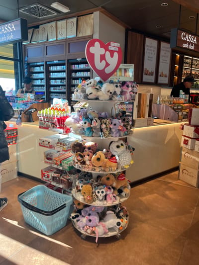The image displays the checkout area of a retail store, likely an Eataly establishment, given the prominent branding, located in Dorno, Italy. The indoor scene is brightly lit by artificial lighting, featuring a warm-toned tiled floor and modern wooden and light-colored counter surfaces.
In the foreground, several clear plastic bins are filled with a vibrant collection of plush toys, predominantly "Beanie Boos" identified by their characteristic "ty" heart-shaped tags. A light blue plastic shopping basket with black handles rests on the floor nearby.
Towards the mid-ground, a "CASSA cash desk" sign, presented in both Italian and English, hangs above a counter. Behind this counter, a person dressed in a dark top is partially visible, attending to the register area which is stocked with various products. Shelves filled with merchandise line the wall behind the counter.
The store's identity is reinforced by multiple "EATALY" brandings visible on boxes and signs. Other Italian texts include "PRODOTTI BUONI DI EATALY" (Good products of Eataly) and what appears to be "CONCEDITI UNA PAUSA DI QUALITÀ" (Treat yourself to a quality break), indicative of a focus on gourmet items and customer experience. A sign also mentions "CON GUARANA," likely an ingredient or product name. The overall scene suggests an active, well-organized retail environment, typical of a specialty food market or a department store section.