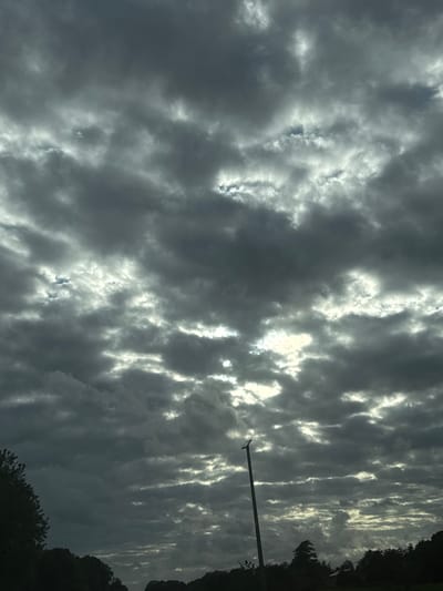 The image shows a cloudy sky over Wierden, Netherlands. The clouds are dark and ominous, with some patches of light breaking through. A lamppost is visible in the lower right portion of the image, and trees and foliage line the bottom of the frame. The overall scene suggests an overcast day with the possibility of rain.