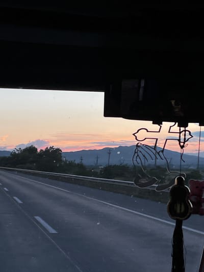 The image shows a view from inside a vehicle, looking out at a highway at sunset. The sky displays soft hues of pink, orange, and yellow. In the foreground, on the right side of the windshield, a religious ornament featuring praying hands and a cross is visible. Below this ornament, a dark, teardrop-shaped charm with a tassel hangs down. A small pink teddy bear is attached to the right side of the ornament. The highway stretches into the distance, with markings on the asphalt. Beyond the highway, there are trees and hills, with power lines visible in the middle ground. The overall scene suggests a journey through the Georgian countryside near Dzveli Kanda as the day ends.