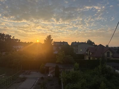 The image captures a sunrise over a suburban area, likely in Marki, Poland, given the architectural styles of the buildings. The sun, a bright orb, is positioned in the left half of the frame, casting a warm, golden light across the sky, which is filled with scattered, textured clouds. Tree lines and building rooftops are silhouetted against the vibrant sky in the background. In the foreground, there are residential buildings with red-tiled roofs and modern apartment blocks. The lower part of the image shows a garden or backyard area with fencing, a paved patio with a picnic table, some greenery, and a small shed or outbuilding. The overall scene depicts a peaceful morning in a residential neighborhood. There are no people visible in the image.