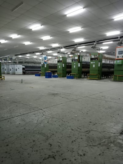 The image displays the interior of a large industrial factory floor, likely a textile or yarn production facility, located in Nglundo, Indonesia. The space is dominated by numerous rows of heavy industrial machinery, predominantly colored olive green and white, extending into the background. These machines appear to be spinning or winding units, fitted with many spools of white and off-white thread or yarn.

The floor is made of concrete, showing significant signs of wear with prominent cracks and various stains or debris scattered across its surface. Scattered intermittently between the machinery are blue plastic crates or baskets, and yellow metal safety barriers or frames are positioned in front of some equipment. Numbers "0", "1", "2", "3", "4", and "5" are clearly visible on the front panels of the green machines, likely serving as identification markers. Control panels are also visible, including a blue unit with an orange indicator light on the left and an orange-faced panel with a digital screen on the right.

The ceiling consists of a grid of light-colored panels, brightly illuminated by numerous long, rectangular fluorescent light fixtures, which are the primary source of light in the facility. There are no people visible in the image, suggesting the machinery is currently idle or the factory is not in active operation. The absence of people and the bright artificial lighting mean the time of day and external weather are indeterminate. The overall scene conveys a functional, albeit currently quiet, industrial environment.