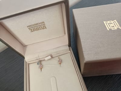A jewelry box containing a pair of rose gold earrings with pink heart-shaped gems and a blue gem is positioned on a dark, textured surface. The interior of the box displays a logo in gold lettering that reads "МОСКОВСКИЙ ЮВЕЛИРНЫЙ ЗАВОД," translating to "Moscow Jewelry Factory." To the right of the open jewelry box is a closed, gray gift box with a gold emblem. The scene appears to be indoors, possibly in a home or retail setting. The location is specified as Первомайское, Russia.