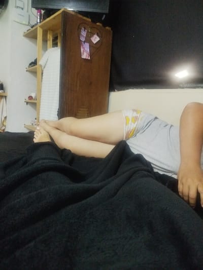 A person is lying on a bed, covered partially by a black blanket. The person is wearing a gray shirt and patterned underwear. The person's feet are visible at the edge of the blanket. In the background, there is a shelf unit and a dark wall or curtain. The location is possibly a bedroom in a residence, based on the furniture and belongings. There is no verifiable evidence to confirm a location in Catia La Mar, Venezuela.