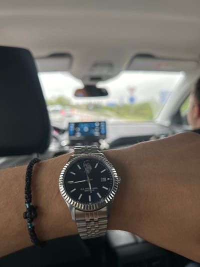 The image captures a close-up perspective from inside a vehicle, focusing on a person's left forearm and wrist. The main subject is a silver-toned wristwatch with a dark blue or black dial, featuring a fluted bezel and a jubilee-style metal bracelet. The watch displays a date window at the 3 o'clock position showing "26". Below the watch, a black braided string bracelet with a small knot adorned with light blue beads is worn on the wrist.

In the blurred background, elements of the car's interior are visible, including what appears to be a dashboard or central console area with a screen showing some illuminated graphics. Through the front windshield, a bright, possibly overcast, daytime sky is discernible, along with hints of green foliage or trees. The perspective suggests the wearer is either a passenger or driver in a car.

Visible text on the watch face includes "U.S. POLO ASSN." and "SINCE 1890", along with the watch's minute markers and the date "26". The overall scene suggests a moment of travel or waiting within a vehicle, located in Mikre, Bulgaria.
