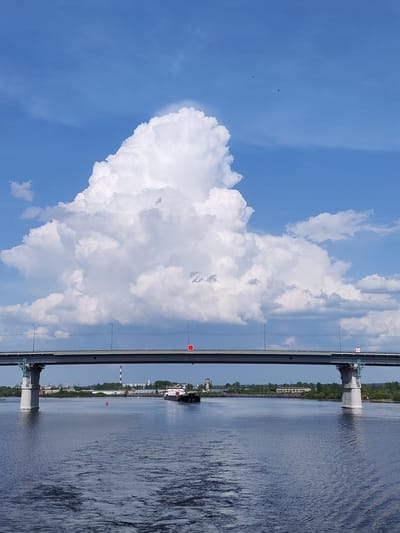 A long, gray bridge spans a wide body of dark, rippled water, with a single barge navigating the waterway beneath. The supports of the bridge are prominent, visible on either side. The bridge itself has lampposts along its length.
A large, fluffy, cumulus cloud dominates the bright blue sky behind the bridge, taking up a significant portion of the upper half of the image.
In the background, the skyline shows a mix of industrial buildings, including a smokestack, and green vegetation along the shoreline. A red buoy is visible in the water in the foreground.
While I cannot confirm that this is the city of Нифантово, Russia, there is nothing in the image to suggest that it is not.