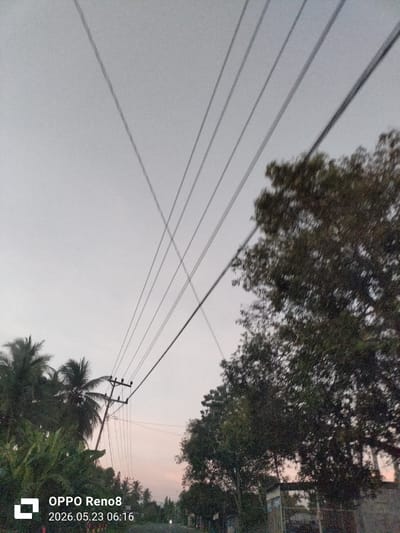 The image depicts an outdoor scene in Gampongrayadagang, Indonesia. In the foreground, power lines stretch across the sky, intersecting near the center. To the left, there are palm trees and various green plants. A utility pole is visible among the palm trees. To the right, there are trees with dense foliage. The sky is overcast, with a hint of faint orange near the horizon. A white rectangular logo with "OPPO Reno8" and the date "2026.05.23 06:16" is located in the lower left corner of the image.