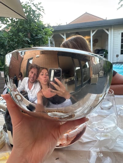 A high-angle, medium shot captures a person holding a large, highly reflective silver bowl in their hand, which is prominently featured in the foreground. The bowl acts as a convex mirror, reflecting a scene occurring behind it. In the reflection, a couple is visible. The male subject, on the left, wears a white open-collared shirt and smiles broadly. The female subject, on the right, is taking the photo with her smartphone. She wears a black tank top and puckers her lips, making a kissy face, while her left hand is raised, holding her phone and displaying a peace sign. A third person is partially visible behind them, out of focus. The background of the reflection shows a brick building, foliage, and what appears to be a swimming pool and outdoor furniture. The surrounding environment, as seen both in and out of the reflection, suggests a residential outdoor setting, possibly a backyard with a pool and a covered patio area. The overall scene conveys a casual, social gathering. The image was taken in the city of Ridderkerk, Netherlands.