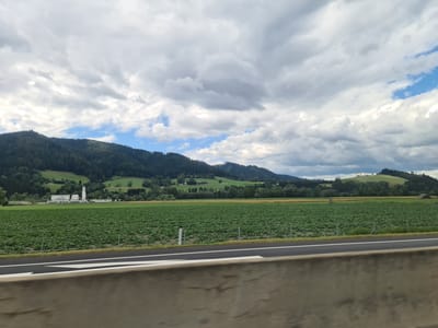 An expansive, wide-angle outdoor photograph captures a scenic rural landscape under a dramatic, cloudy sky. The foreground features a concrete barrier and a paved road with white and black lane markings, suggesting a highway or a similar thoroughfare. Beyond the road, a vast, flat field of lush green crops stretches towards a range of rolling, tree-covered hills in the mid-ground. A cluster of white buildings with tall structures, possibly industrial or agricultural facilities, is visible on the left side of the field. The background is dominated by a chain of densely forested mountains, their peaks partially obscured by the heavy, grey clouds that fill the upper portion of the frame. Patches of blue sky peek through the clouds, hinting at the possibility of clearing weather. The overall impression is one of a tranquil, undeveloped rural area. The location is identified as Kaisersberg, Austria.