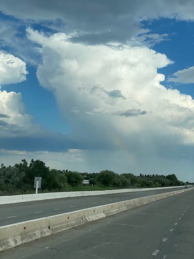 The image depicts a highway scene with a dramatic sky and a faint rainbow. The highway, bordered by concrete barriers, stretches into the distance, with a line of trees and vegetation on the left. In the background, a white RV is visible among the trees. The sky is filled with a mix of bright white cumulus clouds and darker, ominous storm clouds, with a hint of blue sky peeking through. A subtle rainbow arcs across the sky, suggesting recent or ongoing rainfall. The overall impression is one of a dynamic weather event occurring over a rural or semi-rural landscape, possibly a highway on the outskirts of Qtay, Uzbekistan, given the prevalence of highway infrastructure and vegetation. There are no visible people or specific interactions.