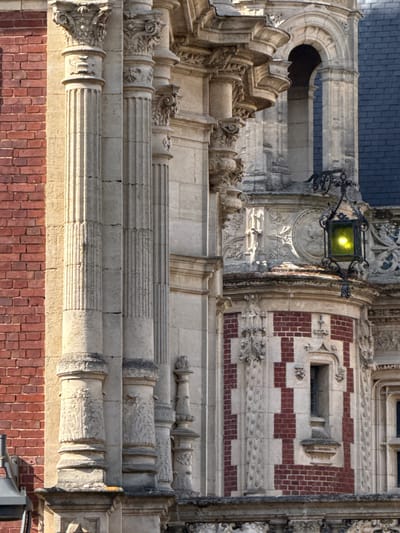 This detailed image captures a section of a highly ornate building facade, presented rotated 90 degrees clockwise. The architecture combines various materials and elaborate decorative elements, indicative of a historical or grand structure, likely located in Fécamp, France.
The top portion of the image displays a dark blue/grey tiled roof. Below this, a grand, arched stone opening, possibly part of a window or a recessed balcony, is visible, featuring intricate carvings and decorative moldings.
Towards the center of the image, an elaborate black metal lamppost is prominently featured, attached to a richly sculpted stone section. The lamppost emits a bright yellow light, even though the scene appears to be set during the daytime, with strong, angled sunlight casting deep shadows across the facade and highlighting its textures. Adjacent to this, the building's surface transitions into a striking pattern of alternating red brick and light-colored stone bands, creating a checkerboard-like design. A small, rectangular window with a stone frame is set within this patterned brickwork.
Further down the facade, multiple levels of decorative stonework are apparent. Several columns and pilasters are visible, showcasing diverse designs; some are fluted, while others are extensively carved with foliage, scrollwork, and what appear to be grotesque figures, characteristic of Gothic or Gothic Revival architectural styles. The stone exhibits signs of age and weathering in various places. The lowest visible section of the building consists of more uniform red brickwork.
No people or explicit activities are depicted in the image. The focus is entirely on the intricate craftsmanship and textural qualities of the building's exterior under clear, sunny conditions. No visible text is present.