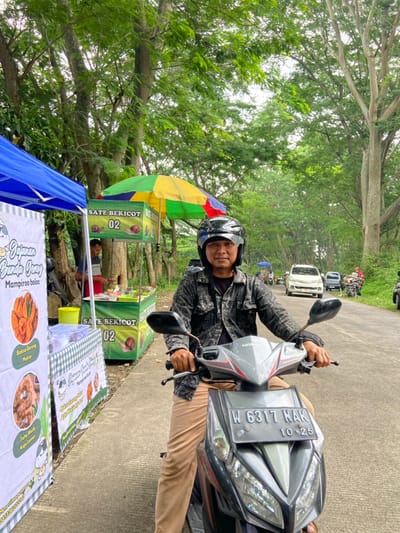 A man on a motorcycle is positioned on a paved road lined with trees. He's wearing a helmet and a jacket. Beside the road, a food stall is set up with a sign advertising "Sate Bekicot 02," and another sign offering "Gajuma Bunda Gamy Mampiroo boloo," along with pictures of food items like "Baksa Goreng" and "Tate Waik." Further down the road, there are other vehicles parked. The motorcycle's license plate reads "W 6317 NAK 10-25."