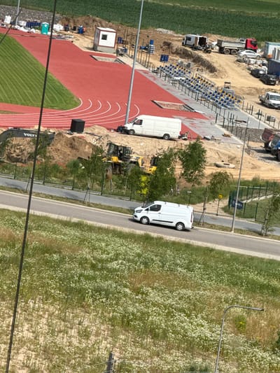 The image captures a construction site with a newly laid red running track and a section of green turf, characteristic of a sports stadium or athletic facility. Several vehicles are present, including a white van driving on an adjacent road, and a white delivery van parked on the construction site. Construction equipment, such as a yellow excavator, is also visible. In the background, bleachers are erected, indicating spectator seating. The overall scene suggests ongoing development or renovation of a public sports venue. The location is identified as Mikołajki, Poland, a town known for its tourist attractions, including sports facilities.
