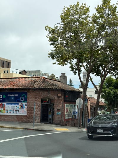 The image shows a street corner in Berkeley, California, United States of America. A black Toyota sedan is parked on the right side of the street. In the background, there is a brick building with a red tile roof. A sign on the building advertises "Seven Palms EBT". Further back, a clock tower is visible. Trees line the street, and the sky is overcast. The overall scene is a typical urban streetscape with a mix of commercial and residential buildings.