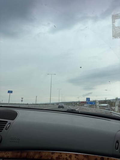 The image shows a view from inside a car, looking out through the windshield. It appears the car is driving on a highway or major road. On the side of the road, there are lampposts and road signs. The sky is overcast. The dashboard of the car is visible in the foreground.
It is difficult to say for sure, but the location is not in the city of Kosh, Armenia.