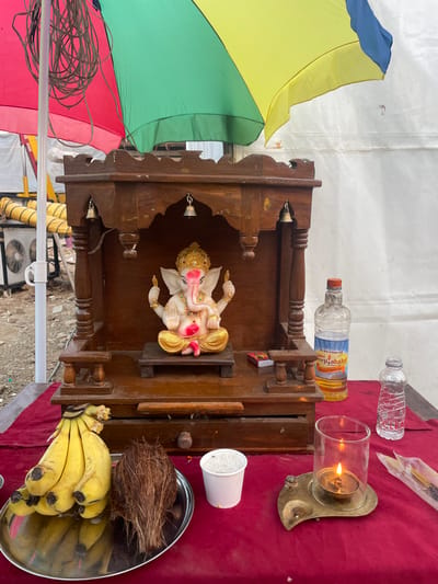 The image shows a Hindu shrine setup outdoors, likely in Mira-Bhayander, India. The shrine is made of dark wood and houses a statue of Ganesha, adorned with gold and pink details. Offerings are arranged around the shrine on a red surface, including bananas, a coconut, a cup of white granular material, and bottles of water and oil, along with a lit candle. To the left of the shrine is a multi-colored umbrella and a coil of wires. An air conditioning unit can be seen in the background. The ground appears to be concrete.