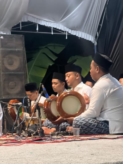 In Wedarijaksa, Indonesia, a group of men wearing black peci hats and white shirts are playing traditional drums. The men are seated on the ground, some are holding the drums, while others are actively playing them. Microphones are positioned in front of the drums to amplify the sound. In the background, large speakers and a white tent with decorative frills are visible. The lighting suggests the scene is taking place at night or in a dimly lit indoor setting. The ground is covered with rugs and cables, indicating a performance or event.