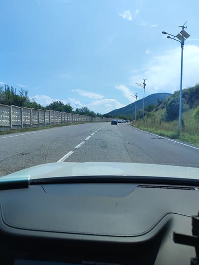 The image shows a view from inside a vehicle driving on a road in Chinarkent, Uzbekistan. The road has white dashed lines marking the lanes. On the left side of the road, there is a long, continuous concrete fence with a decorative pattern. Beyond the fence, trees and vegetation are visible. On the right side of the road, there is a steep, vegetated hill. Several streetlights with solar panels and small wind turbines are spaced along the right side of the road. In the distance, several cars are driving on the road. The sky is blue with scattered white clouds, and the sun appears to be shining. The foreground shows the dashboard of the vehicle.