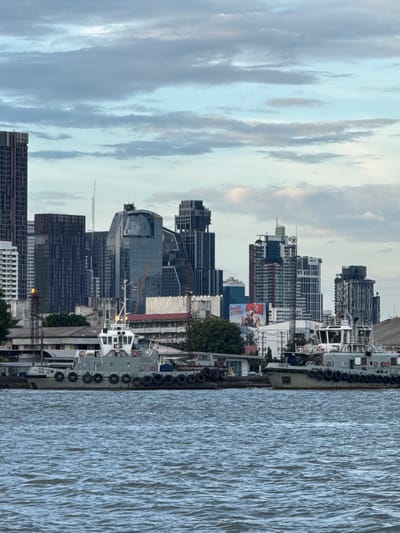 The image shows a waterfront cityscape with modern high-rise buildings in the background and two tugboats in the foreground on a river. The sky is cloudy with some patches of blue. The buildings display a variety of architectural styles, including glass facades and angular designs. The tugboats are grey with white superstructures and appear to be docked or moving slowly. The river water is choppy. A billboard with Thai text and images is visible on one of the buildings. The overall scene suggests an urban port or industrial area. The location is identified as Bang Kachao Subdistrict, Thailand.