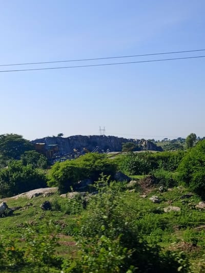 The image shows a quarry site nestled within a hilly, vegetated landscape, under a clear blue sky. In the foreground, lush green bushes and grasses cover the sloping terrain, with scattered rocks. Further back, the terrain rises to form a rocky hill face, where the quarry operations are situated. Several structures, appearing to be part of the quarry infrastructure like buildings and conveyor belts, are visible against the rock face. In the distance, more rolling hills and scattered trees can be seen, with a power pylon visible on the horizon. Two parallel power lines stretch across the upper part of the sky. The scene appears to be located in the rural outskirts of Mara, Nigeria, characterized by its natural topography and industrial activity. There are no people visible in the image.