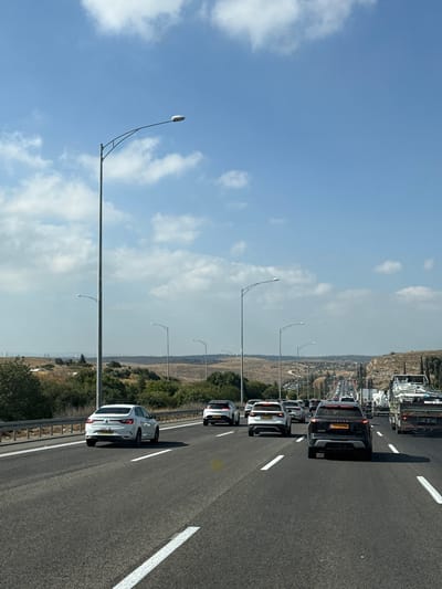 The image depicts a multi-lane highway in Israel, with moderate traffic under a bright, partly cloudy sky. The perspective is from a vehicle moving along the highway, looking ahead. On the right side of the road, a steep, rocky embankment covered with sparse vegetation is visible. Several tall streetlights with modern fixtures line both sides of the highway. In the distance, the road curves and ascends into a hilly landscape, with more vehicles and streetlights visible stretching into the horizon. The foreground shows the asphalt road with white lane markings. Several cars and a large truck are the primary subjects on the road. A white sedan is in the left lane, a white SUV and a black SUV are in the middle lanes, and a truck with a load of white pipes or beams is in the right lane. The overall environment suggests a sunny day in a rural or semi-rural area of Israel.