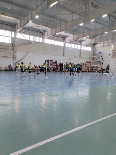 An indoor volleyball game is taking place in a gymnasium in Vinogradny, Russia. Several young female athletes in blue and yellow uniforms are on the court, actively engaged in play. One player in the foreground is in the process of striking a volleyball. Other players are positioned on their side of the net, anticipating the next move. Spectators are seated on benches along the sidelines, observing the match. The gymnasium features a high ceiling with exposed beams and industrial lighting, typical of sports facilities. The court is marked with white lines, and a net divides the playing area. The overall atmosphere suggests a competitive sporting event within a functional, no-frills gymnasium.