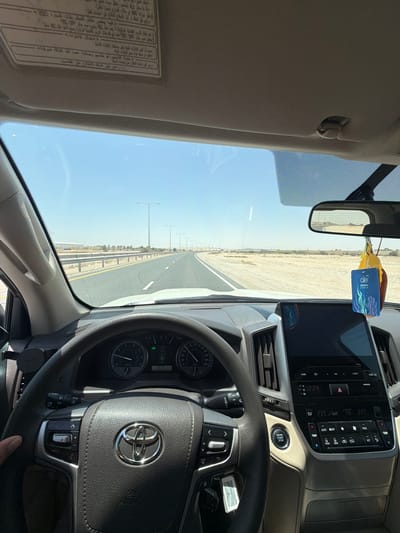 The image provides an interior view of a vehicle, likely a Toyota given the steering wheel logo, driving on a desert road under a clear blue sky. A hand is visible on the steering wheel, indicating a driver.

The dashboard features a large infotainment screen displaying "10:24" and a climate control panel set to "23.5" degrees, with buttons for "ENGINE START STOP," "AUTO," "DUAL," and "A/C." The instrument cluster shows an odometer reading of "88888" and various gauges. An "oer power pocket" air freshener hangs from the rearview mirror. A "PARLIAMENT" cigarette pack is visible near the steering column.

The passenger-side sun visor contains a label with Arabic text, part of which reads: "فيما يلي نظام التعليم باليات السيارة 150 واستقبال الإطارات. دمة التنجيم: أم تتشحيم وصالات عود تقل الطاقة الحركية. ورائع كليب الخدمة الاطلاع على معلومات المسيلة راجع داخل الملك للاطلاع على النامي". This text likely contains instructions or warnings related to the vehicle.

Outside, a straight asphalt road stretches into the distance, marked with white and yellow lines. The road is bordered by guardrails and a barren, sandy landscape characteristic of a desert environment. Streetlights are intermittently spaced along the roadside. The clear, bright sky indicates daytime. The scene evokes a drive through an open, arid region, consistent with the location context of Umm Salal Ali, Qatar.