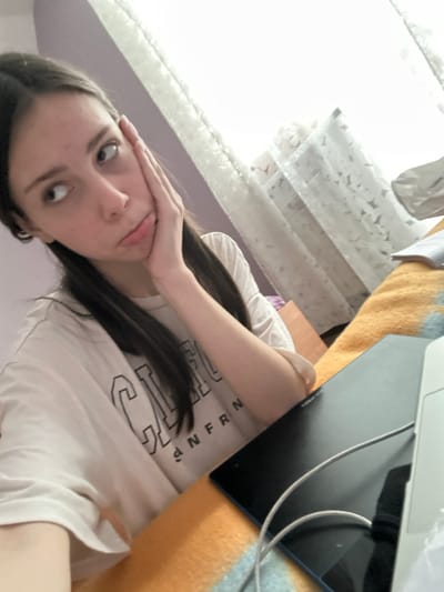 An indoor scene located in Волховец, Russia, features a young adult female seated at a table or desk. The female, appearing to be in her late teens or early twenties, has long, dark brown hair and a fair complexion. She is wearing a light-colored (beige or off-white) short-sleeved t-shirt with dark lettering, partially legible as "CALIFORNIAN". Her right hand is resting against her right cheek, fingers extending towards her temple, and her lips are slightly pursed. She is looking upwards and to the viewer's right.
The table surface in the foreground is covered by a patterned textile, displaying prominent orange/yellow and blue/grey sections. On the table, to the female's right, a black rectangular electronic tablet is visible, alongside a white charging or data cable. A portion of another white electronic device, possibly a laptop or a second tablet, is visible on the far right edge of the frame. The background includes a light purple wall on the left and a window on the right, covered by sheer white curtains with a faint pattern, allowing bright ambient light to enter the room. The exact building or specific area within Волховец is not discernible from the image.