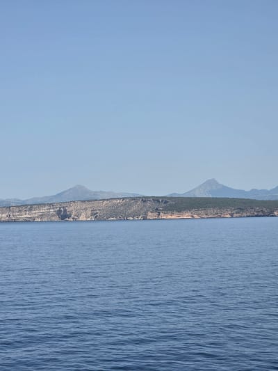 An aerial, medium shot captures the tranquil waters of Sol de Mallorca, Spain, under a clear, pale blue sky. The horizon is dominated by a rugged coastline, characterized by rocky cliffs and a backdrop of distant, muted mountains. The sea, rendered in a deep, rich blue, shows subtle ripples, suggesting a gentle breeze. The overall impression is one of serene natural beauty, with no human presence or man-made structures visible.