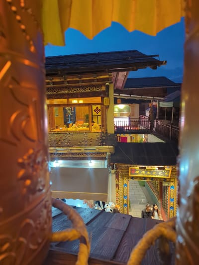 This image captures a nighttime scene in what appears to be a street in Shangri-La, China, viewed through a decorative opening. The perspective is from within a carved, golden structure, possibly a large lantern or architectural element, with rope-like details visible in the foreground.

The scene beyond the opening reveals a narrow street or alley lined with traditional buildings. The architecture features dark wood, intricate carvings, and decorative golden trim with blue accents. Illuminated signs hang from the buildings, one of which prominently displays Chinese characters and the text "Tibetan Embroidery Art of Shangri-La." Lights from within the buildings cast a warm glow on the street, suggesting shops or residences.

In the mid-ground, a few people are visible, including a couple standing on a paved walkway. One person appears to be looking towards the camera. Further inside one of the buildings, a person can be seen through a doorway, illuminated by interior lighting.

The sky above is a deep blue, indicative of twilight or nighttime. The overall atmosphere is one of a vibrant cultural district at dusk, with the illuminated signs and traditional architecture creating an inviting and exotic ambiance. The intricate details of the foreground structure, combined with the cultural signage, strongly suggest this is a tourist-oriented area focused on local crafts and heritage.