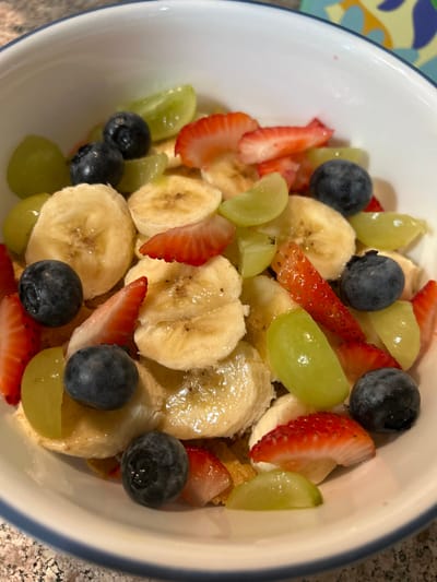 The image depicts a white bowl containing a fruit and cereal mixture. The bowl's contents consist of numerous round slices of pale yellow bananas, various pieces of red sliced strawberries, several whole dark blue blueberries, and multiple light green whole and halved grapes. A clear, glossy liquid appears to coat some of the fruit. Visible beneath the fruit are light brown cereal flakes. The bowl itself features a thin dark blue band along its rim. It is positioned on a speckled, light-colored surface, likely a countertop. In the upper right portion of the background, a fragment of a colorful object with abstract yellow, blue, and orange patterns is visible. No individuals are present, and no actions or interactions are depicted. This scene is located in Thousand Oaks, United States.