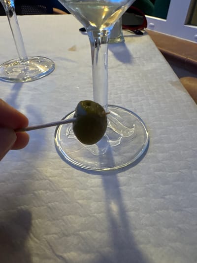 A close-up, eye-level shot focuses on a hand holding a toothpick with a single green olive on it, positioned over the clear glass base of a martini glass. The olive is dark green and slightly wrinkled, with the toothpick inserted through its center. The glass base is circular and transparent, with subtle decorative etchings visible. In the background, another martini glass stem is visible to the left, and a portion of a martini glass filled with a pale yellow liquid is to the right. The surface beneath the glasses appears to be a textured, white tablecloth. The scene is indoors, likely at a restaurant or bar, though no specific city or location is identifiable from the image alone. The lighting is soft, creating some reflections and shadows on the table and glassware.