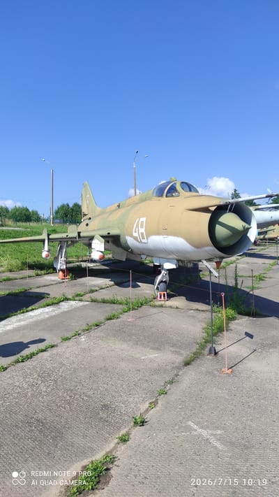 This image depicts an outdoor display of a Sukhoi Su-17 fighter jet, painted in camouflage colors of green and tan, with the number "48" in white on its fuselage. The aircraft is parked on a concrete tarmac with grass growing through cracks. Two streetlights are visible in the background, along with a line of trees and a blue sky with scattered clouds. The scene appears to be a museum or memorial site. The location is identified as Lashetski Sel'ski Sovet, Belarus. The image is timestamped 2026/7/15 10:19. There are no people visible in the frame. The jet has an orange pylon attached to its wing, and a missile or bomb is mounted on it. Another aircraft is partially visible in the background.