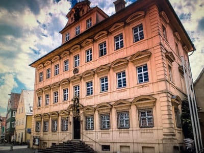 The image shows a historic building in the city of Ellwangen, Germany, specifically the Rathaus (City Hall). The building is a large, multi-story structure with a light peach-colored facade. It features numerous windows with white panes, arranged symmetrically across the different levels. The roof is steep and tiled, with dormer windows and a prominent tower element. The building's architecture suggests a Baroque or Rococo style, with decorative elements above the windows and a central entrance adorned with a statue or sculptural feature. In front of the building, there are stone steps leading up to the main entrance, and a street or plaza is visible in the foreground. The sky above is partly cloudy, with blue sky and white clouds. To the left of the main building, other older buildings with pitched roofs and colorful facades are visible, indicating a historical urban setting. The overall impression is of a well-preserved historical landmark in a European city.