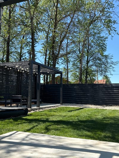 A horizontal shot shows a backyard with trees, a fence, grass, a concrete object, and a wooden structure. The trees are in the left side of the frame, and their branches extend up towards the blue sky. A black wooden fence is to the right of the trees, with a narrow strip of light-colored gravel at its base. Next to the fence is a grassy area. A partially visible concrete object is in the bottom right corner. A black wooden structure with a slanted roof is in the center. It has a series of small, white lights strung along its roofline. The location is a backyard in Юг, Russia.