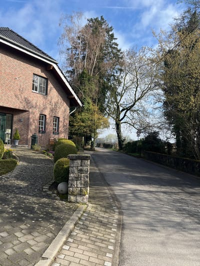 This image captures a street scene in Würselen, Germany, under a partly cloudy blue sky exhibiting an aircraft contrail. Long shadows are cast, indicating direct sunlight from the left.
On the left side of the frame, a two-story residence constructed of red brick is partially visible. It features a dark tiled roof with a gutter system, multiple white-framed windows, and a green front door. A paved walkway, composed of interlocking grey pavers, curves towards the house entrance. This pathway is bordered by several meticulously pruned spherical green shrubs and a stone pillar, built from rectangular blocks, partially covered in moss, with a grey spherical object resting at its base. A black metal bin or planter is positioned near the front door, and a small wooden bench is visible under a window.
To the right, an asphalt road curves gently towards the right and recedes into the distance. A paved sidewalk, constructed from smaller grey pavers, runs alongside the road, separated by a concrete curb. The road and sidewalk are devoid of people or vehicles.
Various trees populate the scene, including tall conifers and deciduous trees, many displaying bare branches consistent with late winter or early spring. Some trees show nascent green foliage. A prominent deciduous tree with a bare canopy stands near the center of the frame, alongside the road. Further down the road on the right, a circular traffic sign with a red border and white center is mounted on a pole, partially obscured by bare tree branches. A low stone wall or dense hedge defines the right edge of the roadway in the mid-ground.