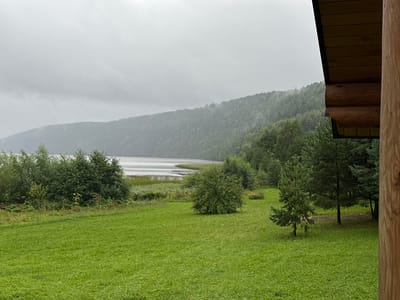 This is a landscape photograph taken from a wooden structure overlooking a grassy field, a lake, and a forested hill under an overcast sky. The scene is set in Berdyash, Russia. In the foreground, a well-maintained green lawn stretches across the frame. To the left and middle ground, various trees and dense foliage are visible, with some taller trees bordering a body of water. The water, likely a lake or a wide river, occupies the middle ground, with wooden structures, possibly docks or piers, extending into it. Beyond the water, a densely forested hill rises, its contours softened by a hazy, possibly rainy, atmosphere. The sky is uniformly gray and cloudy, suggesting a muted or inclement weather condition. A portion of the wooden structure, a log cabin or a similar building, is visible on the right side of the image, its rough-hewn logs and wooden roof providing a rustic foreground element. No people are present in the scene, and the overall impression is one of natural tranquility and perhaps a touch of melancholy due to the weather.