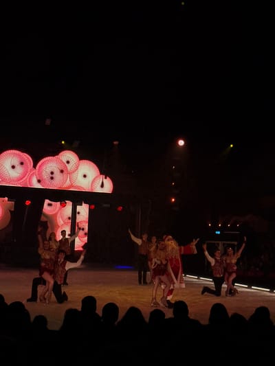 Figure skaters perform on an ice rink illuminated with spotlights and decorative light displays. Pairs of skaters execute lifts and poses, while spectators watch from the sidelines. The background includes geometric patterns and circular lights. The overall atmosphere suggests a festive or theatrical performance.