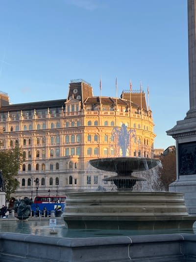 The image displays a daytime scene in Trafalgar Square, City of Westminster, United Kingdom, under a clear blue sky with faint contrails. Golden sunlight illuminates the right side of the frame and the foreground elements.
In the mid-ground, a multi-tiered stone fountain, constructed from a dark material, actively sprays water upwards from its central column and cascades water into its lower basins. To the immediate right, the lower section and carved stone base of Nelson's Column are visible.
In the background, a large, ornate building with a light-colored stone facade, identified as Grand Buildings, is brightly lit by the sunlight. This multi-story structure features numerous arched windows and a dark mansard roof adorned with several flags, including British Union Flags, atop flagpoles.
On the far left, behind the fountain, indistinct figures of people are visible near another sculptural element. A partially visible red double-decker bus is also present behind the people and the fountain. A small "No entry" sign is visible within the foreground basin of the fountain. Sparse tree foliage is discernible in the distant background.