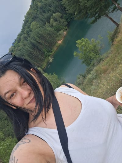 A woman is taking a selfie outdoors with a body of water and a forested hillside in the background. She has long, dark hair that is partially covering her face, and she is wearing a white tank top. Her right arm is visible, showing a tattoo on her shoulder. She is holding a disposable cup in her left hand. The scene appears to be a natural landscape, possibly a lake or quarry, surrounded by trees. The water is a deep teal color, and the hillside is covered in dense green trees. The sky is overcast, suggesting a cloudy day. The time of day is not explicitly indicated, but the lighting suggests it could be late afternoon. The woman's expression is relaxed and content. The location is identified as Dobrotino, Bulgaria.
